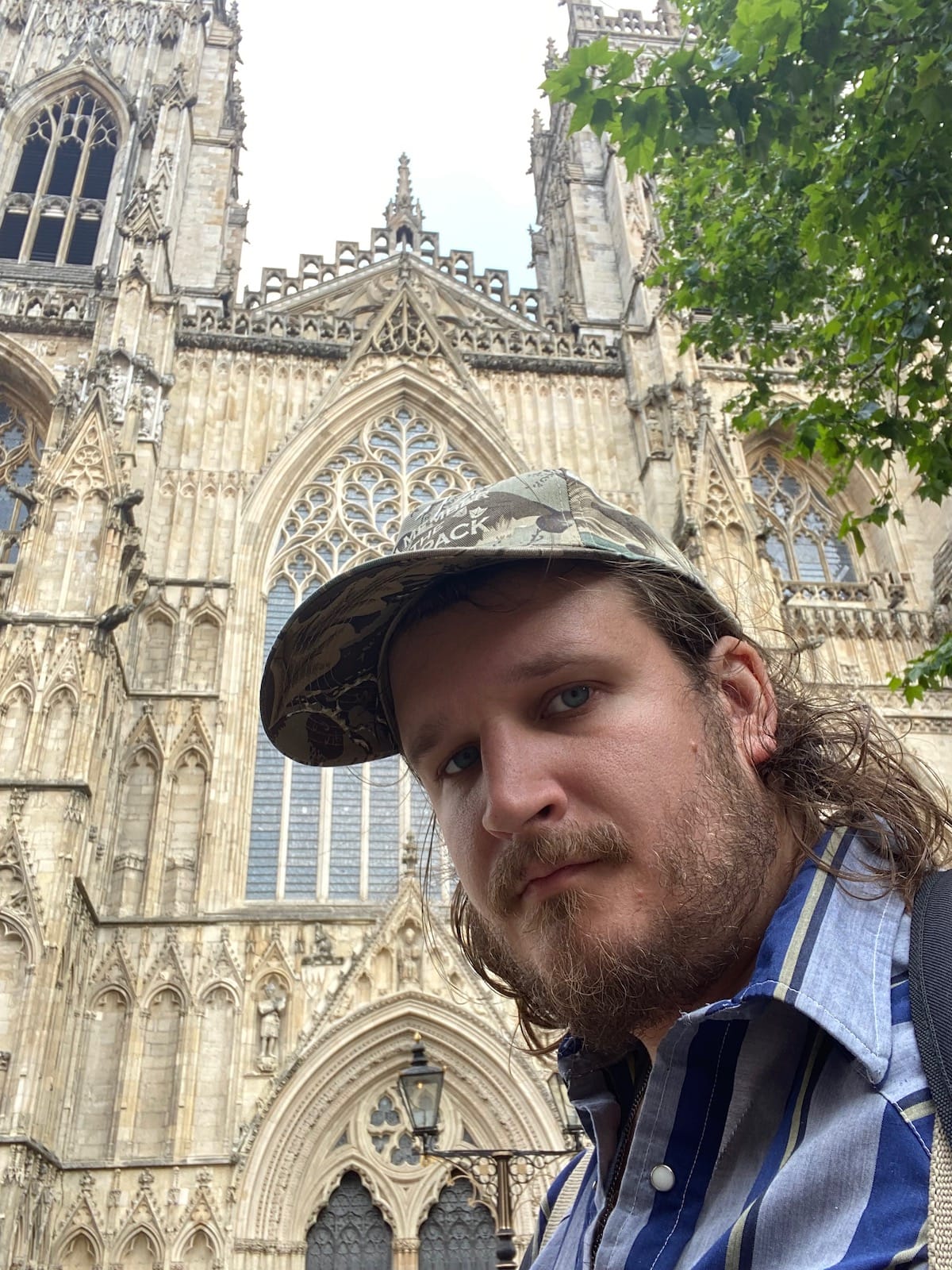 Willi Carlisle in a ball cap stands in front of an old church in England