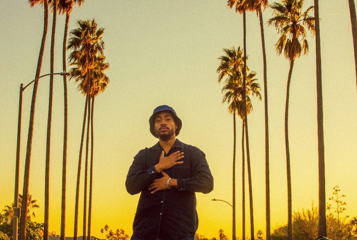 Cadence Weapon against a yellow sky lined with palm trees