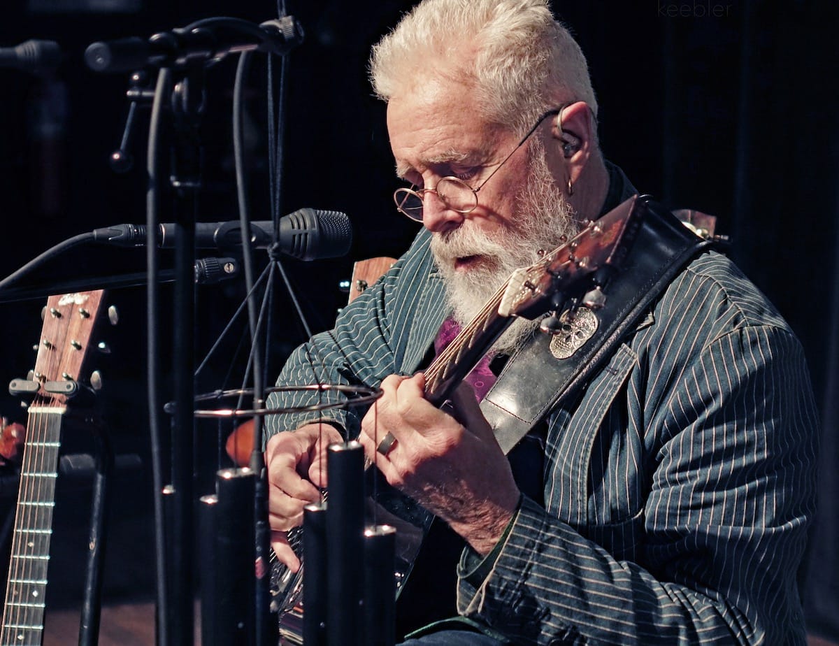 Bruce Cockburn in striped jacked plays guitar onstage