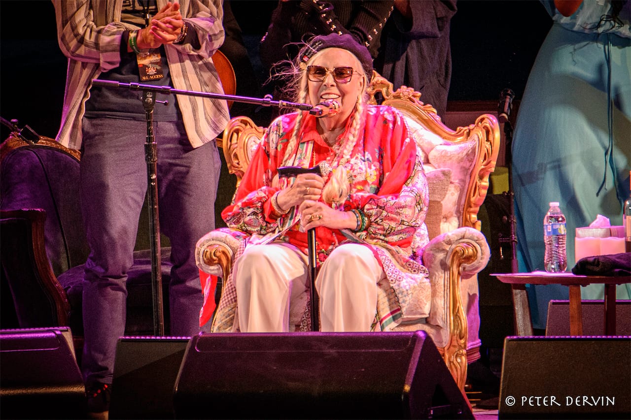 Joni Mitchell performs sitting in an upholstered chair on stage